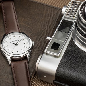 Puede incluir: Un reloj de pulsera plateado con una esfera blanca y una correa de cuero marr&oacute;n. La esfera del reloj tiene marcadores de hora romanos negros y la palabra "WINDSOR" impresa en negro. El reloj est&aacute; sobre una superficie de cuero marr&oacute;n junto a una c&aacute;mara plateada y negra.