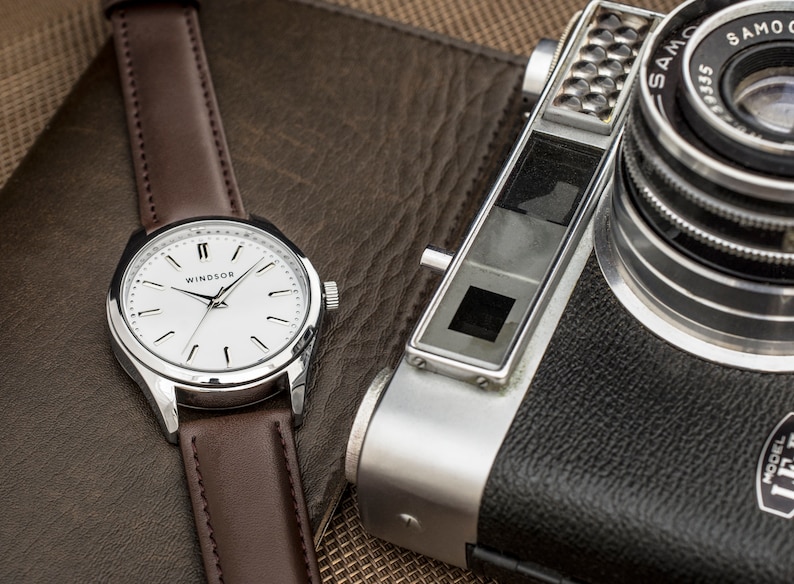 Puede incluir: Un reloj de pulsera plateado con una esfera blanca y una correa de cuero marr&oacute;n. La esfera del reloj tiene marcadores de hora romanos negros y la palabra "WINDSOR" impresa en negro. El reloj est&aacute; sobre una superficie de cuero marr&oacute;n junto a una c&aacute;mara plateada y negra.