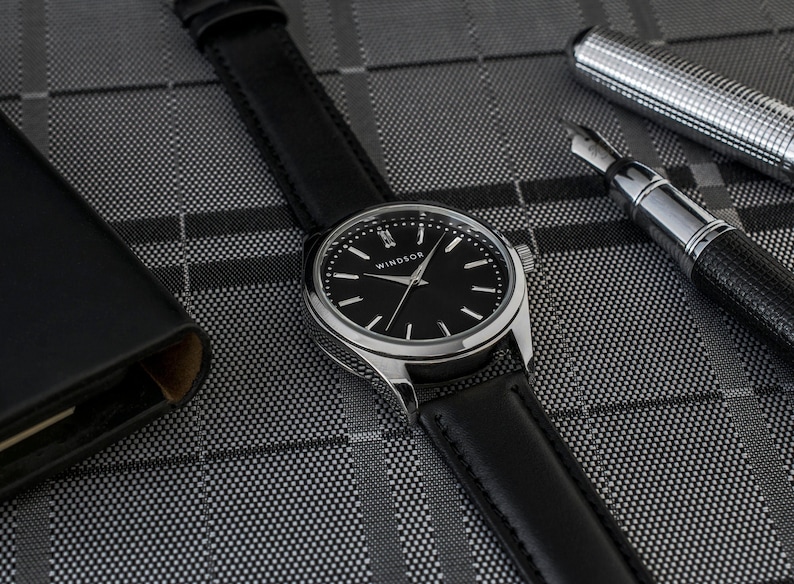 May include: A silver watch with a black leather band and a black face. The watch face has silver hands and silver Roman numeral hour markers. The watch face also has the word "Windsor" printed on it.