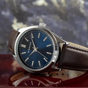 May include: A silver watch with a blue face and brown leather band. The watch face has silver hands and markers and the word "WINDSOR" printed on it.
