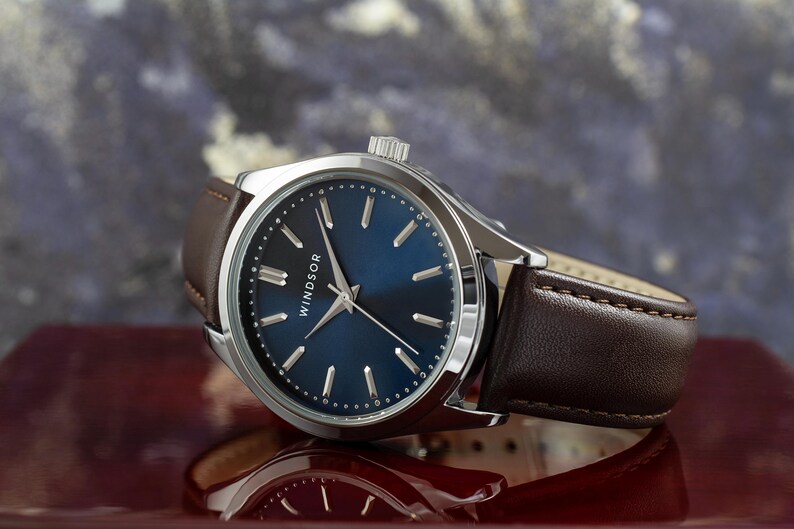 May include: A silver watch with a blue face and brown leather band. The watch face has silver hands and markers and the word "WINDSOR" printed on it.
