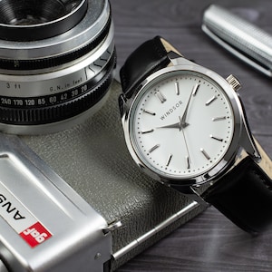 May include: A silver watch with a white face and black leather band. The watch face has black hour and minute hands and black Roman numeral hour markers. The watch face also has the word "WINDSOR" printed on it.