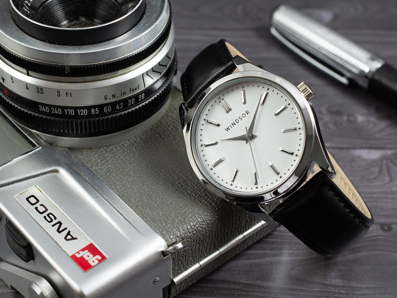 May include: A silver watch with a white face and black leather band. The watch face has black hour and minute hands and black Roman numeral hour markers. The watch face also has the word "WINDSOR" printed on it.
