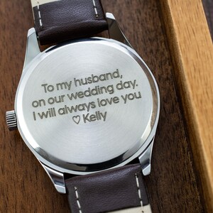 May include: A silver watch with a brown leather band. The back of the watch is engraved with the message "To my husband, on our wedding day. I will always love you ♡ Kelly".