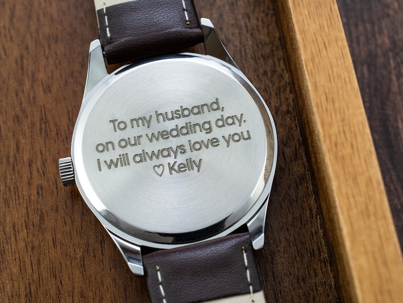 May include: A silver watch with a brown leather band. The back of the watch is engraved with the message "To my husband, on our wedding day. I will always love you ♡ Kelly".