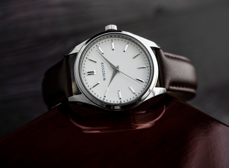 May include: A silver wristwatch with a white face and black Roman numeral hour markers. The watch has a brown leather band and says "Windsor" on the face.