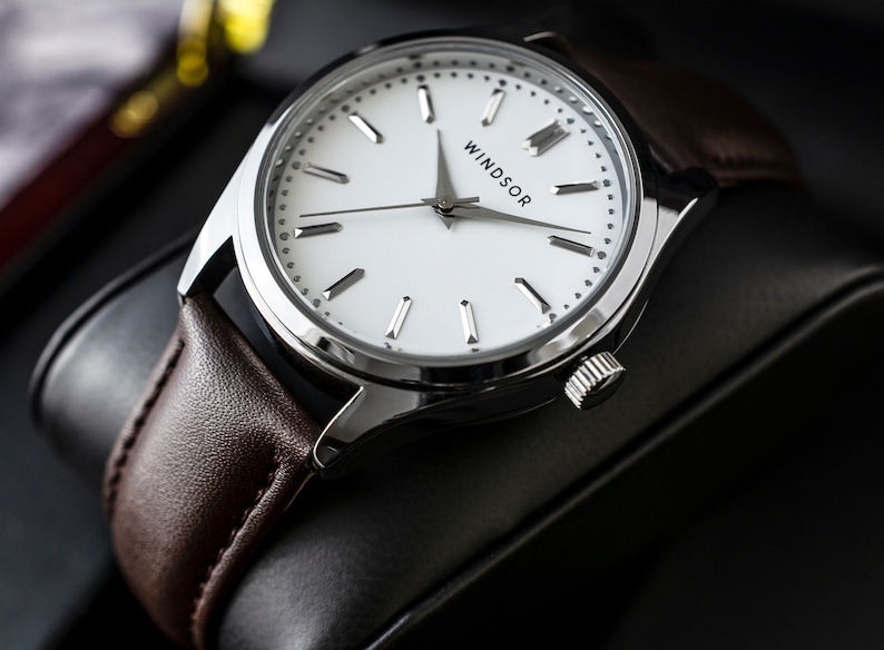 May include: A silver watch with a white face and brown leather band. The watch face has silver hands and hour markers, and the word "WINDSOR" is printed at the top.