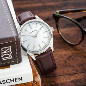 May include: A silver watch with a white face and brown leather band. The watch face has the word "WINDSOR" printed on it. The watch is sitting on a book with the word "TASCHEN" printed on it.