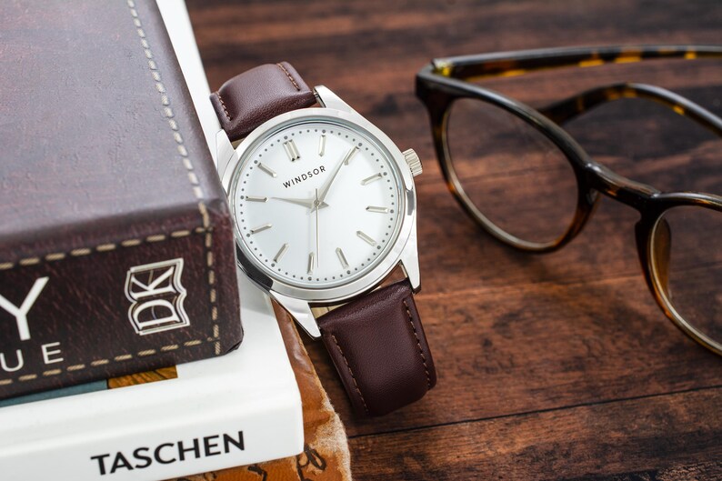 May include: A silver watch with a white face and brown leather band. The watch face has the word "WINDSOR" printed on it. The watch is sitting on a book with the word "TASCHEN" printed on it.