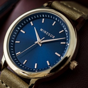 May include: A gold-tone watch with a blue face and rose gold hands. The watch has a brown leather strap and the word "WINDSOR" printed on the face.