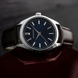 May include: A silver watch with a blue face and brown leather band. The watch face has silver hands and Roman numeral markings. The watch face also has the word "WINDSOR" printed on it.