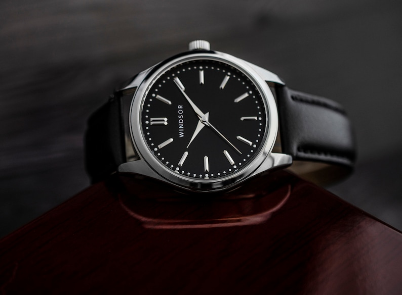 Puede incluir: Un reloj de pulsera plateado con una correa de cuero negro y una esfera negra con marcadores de hora en n&uacute;meros romanos blancos y agujas plateadas. La esfera del reloj tambi&eacute;n tiene la palabra "WINDSOR" impresa.