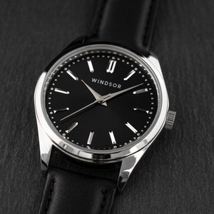 Puede incluir: Primer plano de un reloj Windsor con esfera negra y bisel plateado. El reloj tiene una correa de cuero negro. La esfera tiene marcadores de hora plateados y la palabra "WINDSOR" est&aacute; impresa en blanco.