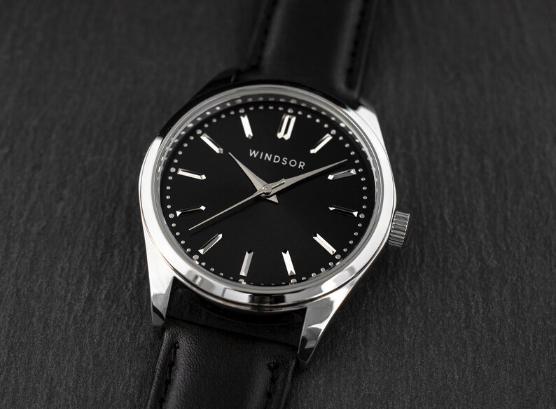 Puede incluir: Primer plano de un reloj Windsor con esfera negra y bisel plateado. El reloj tiene una correa de cuero negro. La esfera tiene marcadores de hora plateados y la palabra "WINDSOR" est&aacute; impresa en blanco.