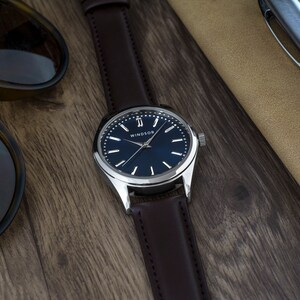 Puede incluir: Un reloj plateado con una correa de cuero marr&oacute;n y una esfera azul. La esfera del reloj tiene agujas plateadas y marcadores de hora en n&uacute;meros romanos. El nombre de la marca "WINDSOR" est&aacute; impreso en la esfera del reloj.