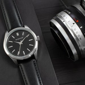 May include: A silver watch with a black leather band and a black face. The watch face has silver hands and Roman numeral hour markers. The watch face also has the word "WINDSOR" printed on it.