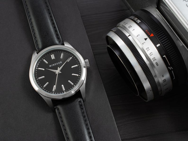 May include: A silver watch with a black leather band and a black face. The watch face has silver hands and Roman numeral hour markers. The watch face also has the word "WINDSOR" printed on it.