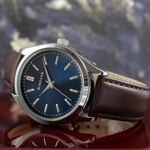 May include: A silver watch with a blue face and brown leather band. The watch face has silver hands and markers and the word "WINDSOR" printed on it.