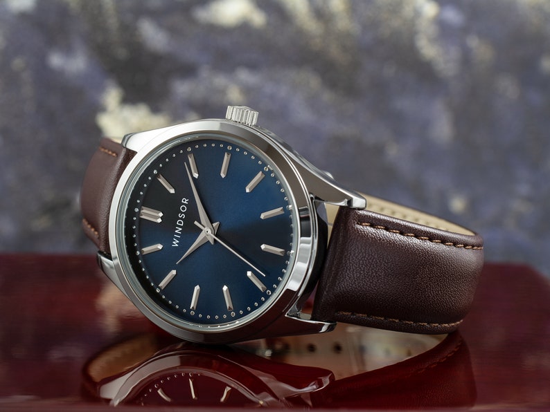 May include: A silver watch with a blue face and brown leather band. The watch face has silver hands and markers and the word "WINDSOR" printed on it.