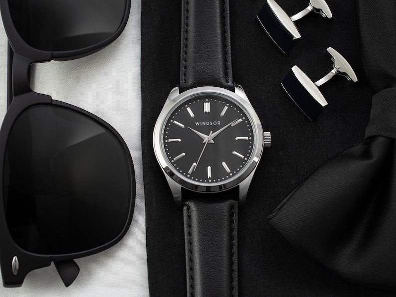 May include: A black leather watch with a silver face and black hands. The watch face has the word "Windsor" printed on it. The watch is on a black surface with cufflinks.