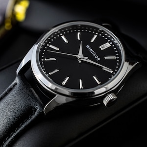 May include: A black leather wristwatch with a silver case and a black face. The watch face has silver hands and hour markers, and the word "WINDSOR" is printed in silver at the bottom of the face.