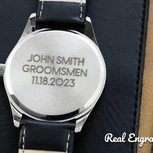 May include: A silver watch with a black leather band. The back of the watch is engraved with the text "John Smith Groomsmen 11.18.2023".