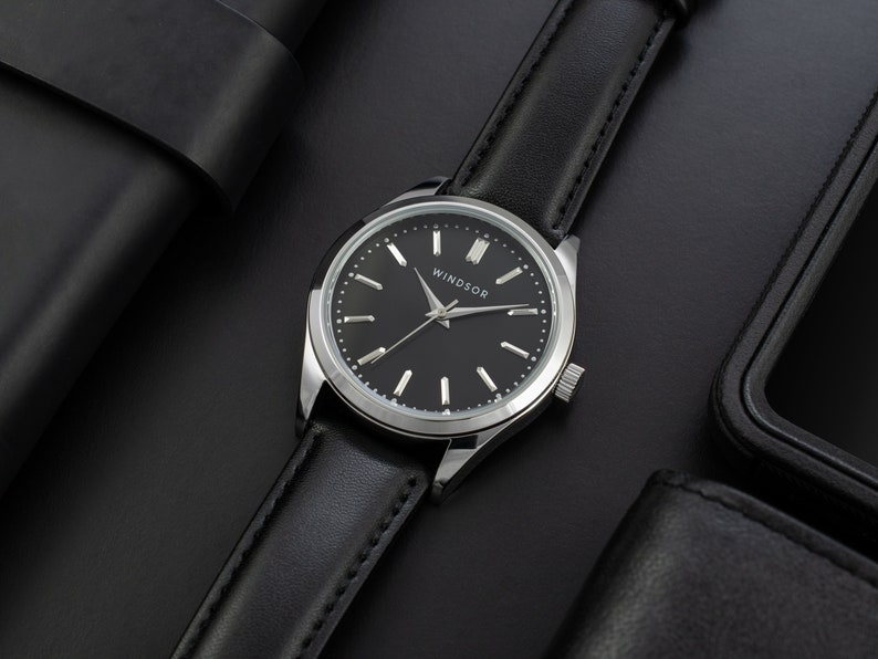 May include: A silver watch with a black leather band and a black face. The watch face has silver hands and markers and the word "WINDSOR" printed on it.