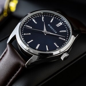 Puede incluir: Primer plano de un reloj Windsor con esfera azul oscuro, manecillas y caja plateadas. La correa es de cuero marr&oacute;n. La palabra "WINDSOR" est&aacute; impresa en blanco en la esfera.