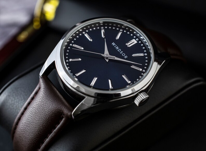 Puede incluir: Primer plano de un reloj Windsor con esfera azul oscuro, manecillas y caja plateadas. La correa es de cuero marr&oacute;n. La palabra "WINDSOR" est&aacute; impresa en blanco en la esfera.