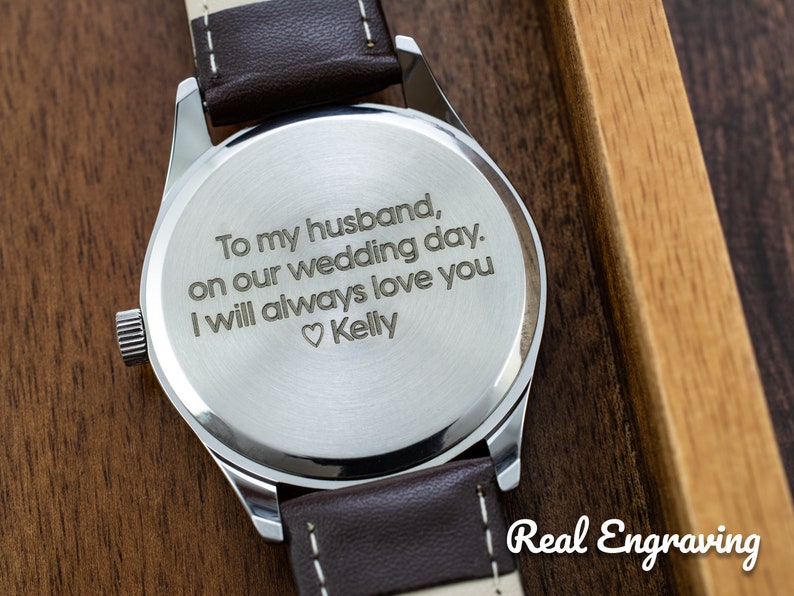 May include: A silver watch with a brown leather band. The back of the watch is engraved with the message "To my husband, on our wedding day. I will always love you ♡Kelly".
