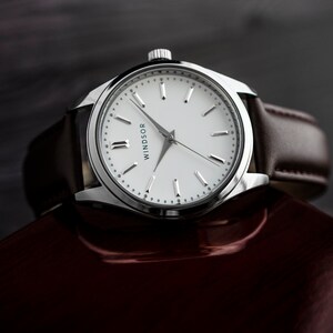 Puede incluir: Un reloj Windsor plateado con esfera blanca y correa de cuero marr&oacute;n. El reloj tiene un dise&ntilde;o minimalista con manecillas y marcadores plateados. El reloj se muestra sobre una superficie roja oscura.