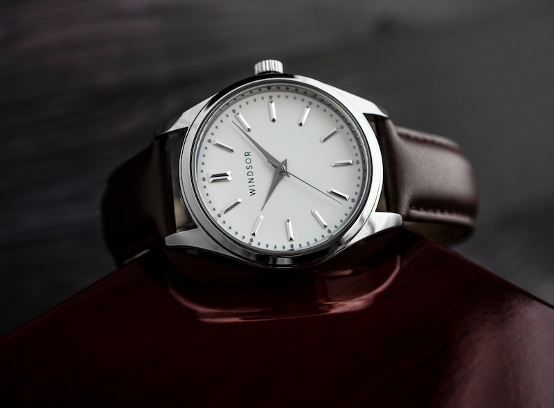 Puede incluir: Un reloj Windsor plateado con esfera blanca y correa de cuero marr&oacute;n. El reloj tiene un dise&ntilde;o minimalista con manecillas y marcadores plateados. El reloj se muestra sobre una superficie roja oscura.