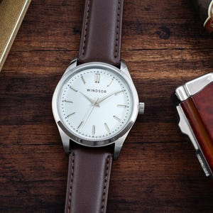 May include: A silver wristwatch with a white face and brown leather band. The watch face has black Roman numeral hour markers and the word "WINDSOR" printed in black.