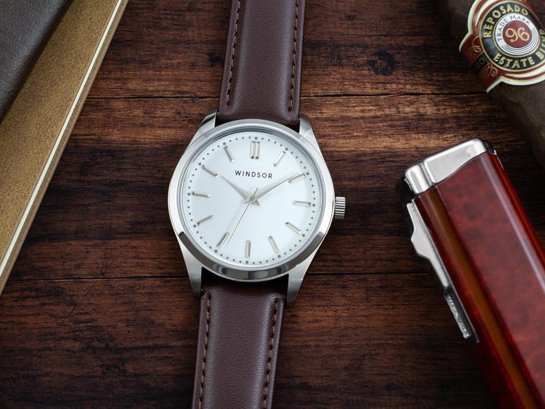 May include: A silver wristwatch with a white face and brown leather band. The watch face has black Roman numeral hour markers and the word "WINDSOR" printed in black.