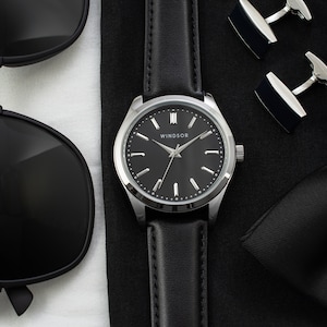 May include: A black leather wristwatch with a silver face and black hands. The watch face has the word "WINDSOR" printed on it. The watch is on a black surface with a black bow tie and black sunglasses.