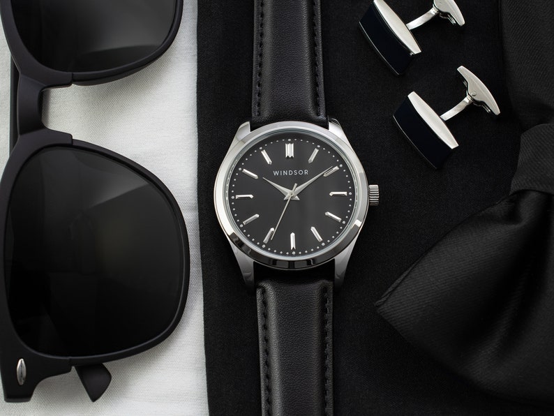 May include: A black leather wristwatch with a silver face and black hands. The watch face has the word "WINDSOR" printed on it. The watch is on a black surface with a black bow tie and black sunglasses.