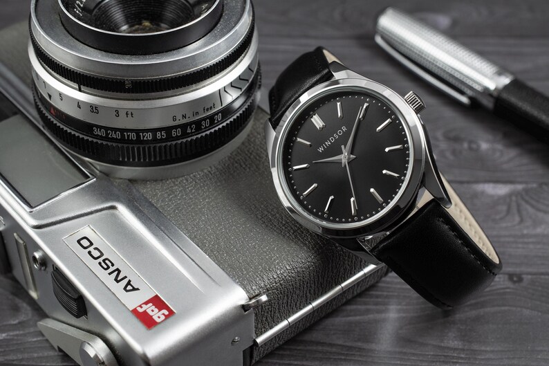May include: A silver watch with a black face and black leather band. The watch face has silver hands and Roman numeral hour markers. The watch face also has the word "WINDSOR" printed on it. The watch is sitting on top of a silver camera.