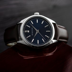 Puede incluir: Un reloj plateado con una esfera azul oscuro y agujas blancas. El reloj tiene una correa de cuero marr&oacute;n y la palabra "WINDSOR" est&aacute; impresa en la esfera.