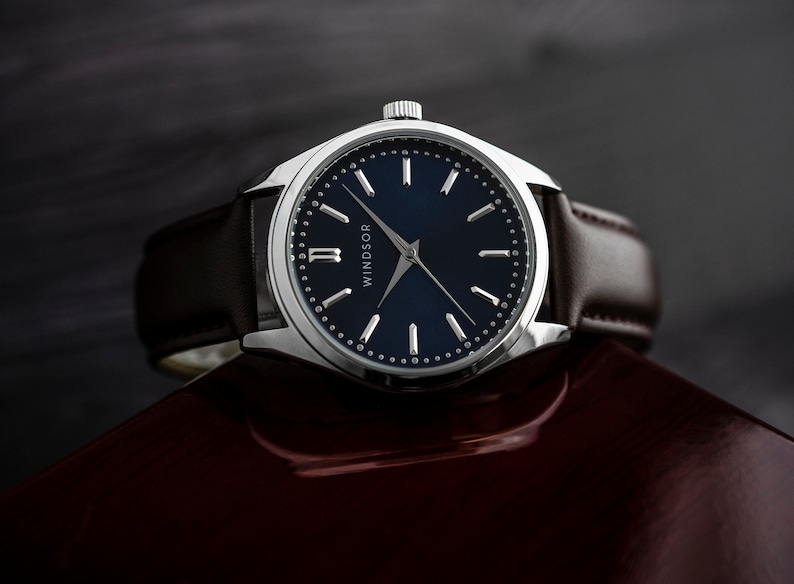 Puede incluir: Un reloj plateado con una esfera azul oscuro y agujas blancas. El reloj tiene una correa de cuero marr&oacute;n y la palabra "WINDSOR" est&aacute; impresa en la esfera.