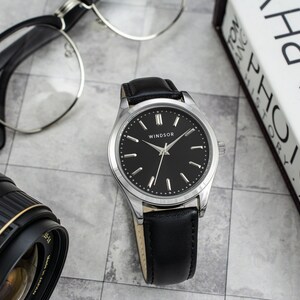 May include: A silver and black wristwatch with a black leather band. The watch face has a black background with silver hands and hour markers. The word "WINDSOR" is printed in silver on the watch face.