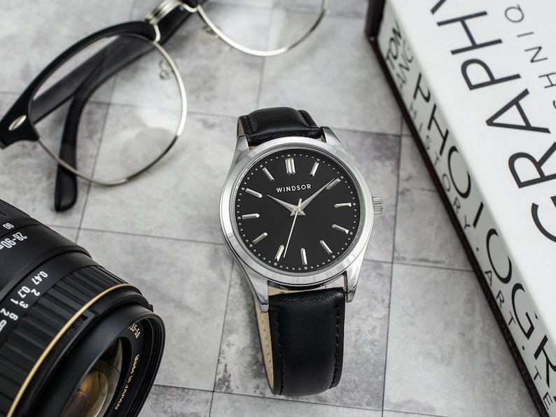 May include: A silver and black wristwatch with a black leather band. The watch face has a black background with silver hands and hour markers. The word "WINDSOR" is printed in silver on the watch face.