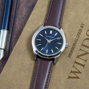 May include: A silver watch with a blue face and brown leather band. The watch face has silver hands and markers. The watch is on a brown surface with the text "Personalized by Windsor Watch Company" printed on it.