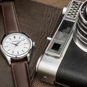 May include: A silver wristwatch with a white face and brown leather band. The watch face has black Roman numeral hour markers and the word "WINDSOR" printed in black. The watch is lying on a brown leather surface next to a silver and black camera.