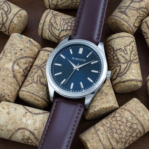 May include: A silver watch with a blue face and brown leather band. The watch face has silver hands and markers, and the word "WINDSOR" is printed in black at the top. The watch is surrounded by wine corks.