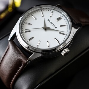 Puede incluir: Primer plano de un reloj de pulsera con esfera blanca y caja plateada. La correa es de cuero marr&oacute;n. La palabra "WINDSOR" est&aacute; impresa en la esfera. El reloj se muestra sobre una superficie negra.