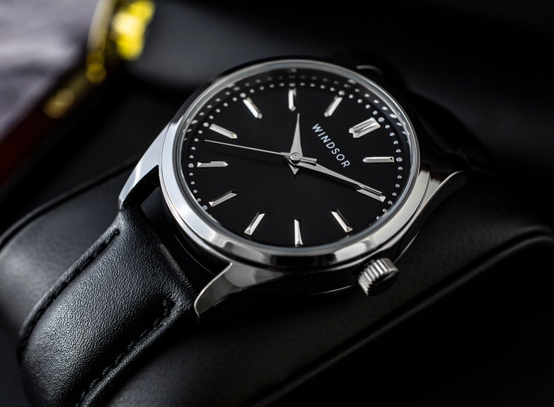 May include: A black leather wristwatch with a silver case and a black face. The watch face has silver hands and hour markers, and the word "WINDSOR" is printed in silver at the bottom of the face.