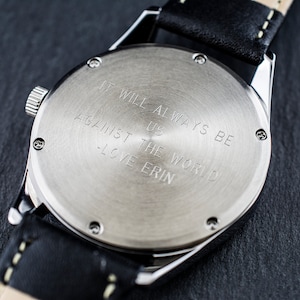 May include: The back of a silver watch with a black leather band. The watch face has the inscription "It will always be against us - the world - love Erin".