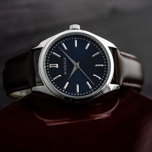 May include: A silver watch with a blue face and brown leather band. The watch face has silver hands and Roman numeral markings. The watch face also has the word "WINDSOR" printed on it.