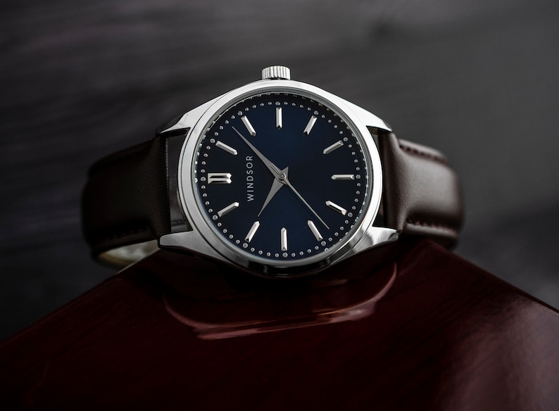 May include: A silver watch with a blue face and brown leather band. The watch face has silver hands and Roman numeral markings. The watch face also has the word "WINDSOR" printed on it.