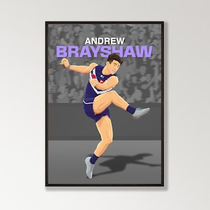 May include: A framed art print depicting a football player kicking a ball. The artwork includes the name "ANDREW BRAYSHAW" in purple text. The player is wearing a blue and white uniform. The print is displayed on a white wall.